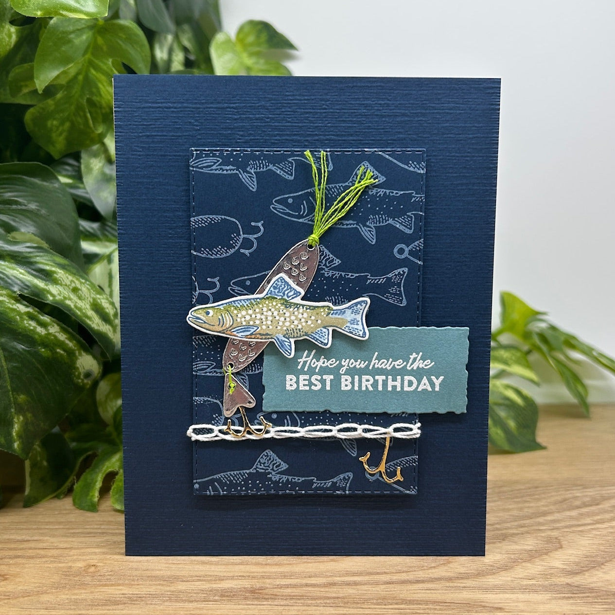 Navy blue birthday card with fish illustration on a wooden surface with green plants in the background