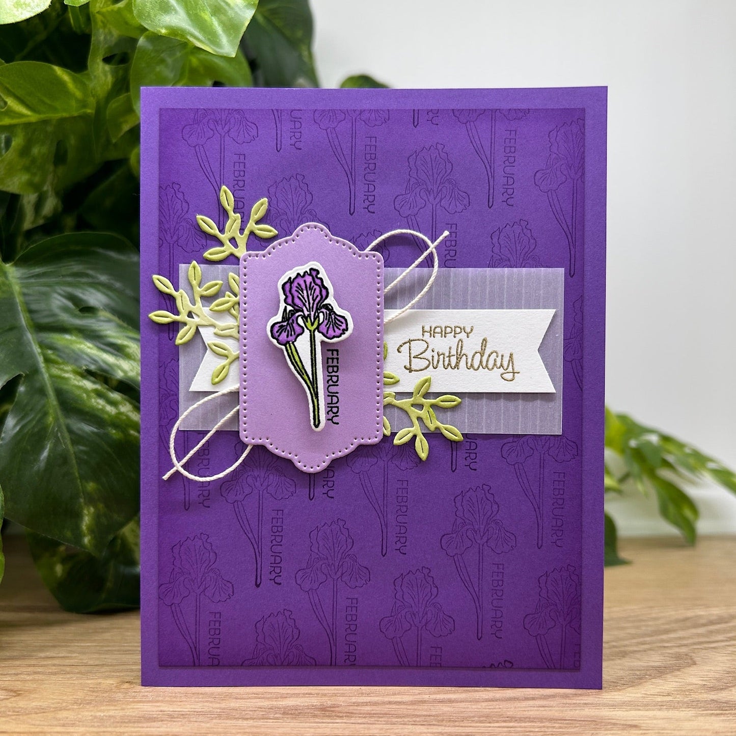 Purple birthday card with floral design and 'Happy Birthday' text, placed on a wooden surface with green plants in the background.