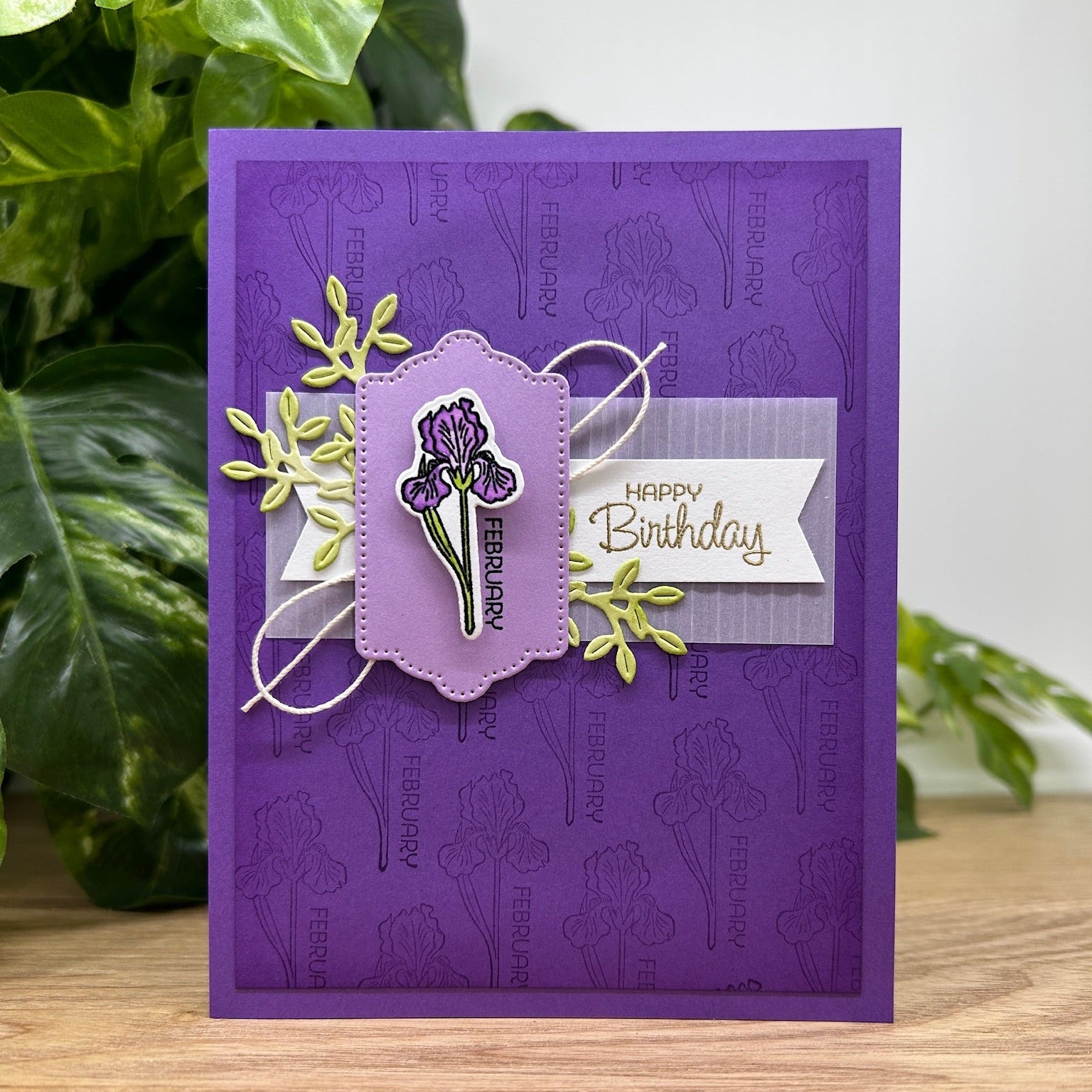 Purple birthday card with floral design and 'Happy Birthday' text, placed on a wooden surface with green plants in the background.
