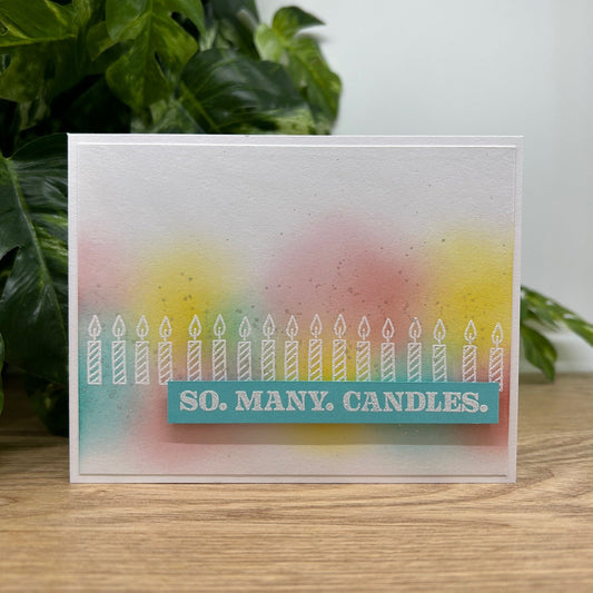 Greeting card with 'So Many Candles' text and candle illustrations on a wooden surface with greenery in the background.