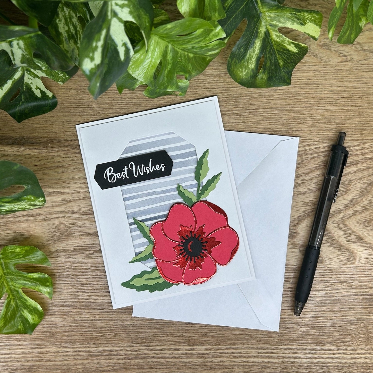 Greeting card with red flower design on a wooden surface with leaves and a pen.