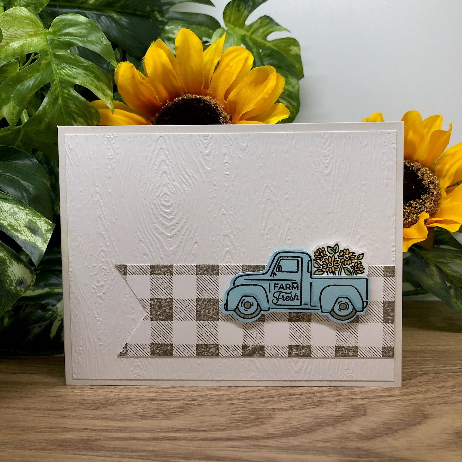 Card with a blue truck and sunflowers on a wooden surface with greenery in the background