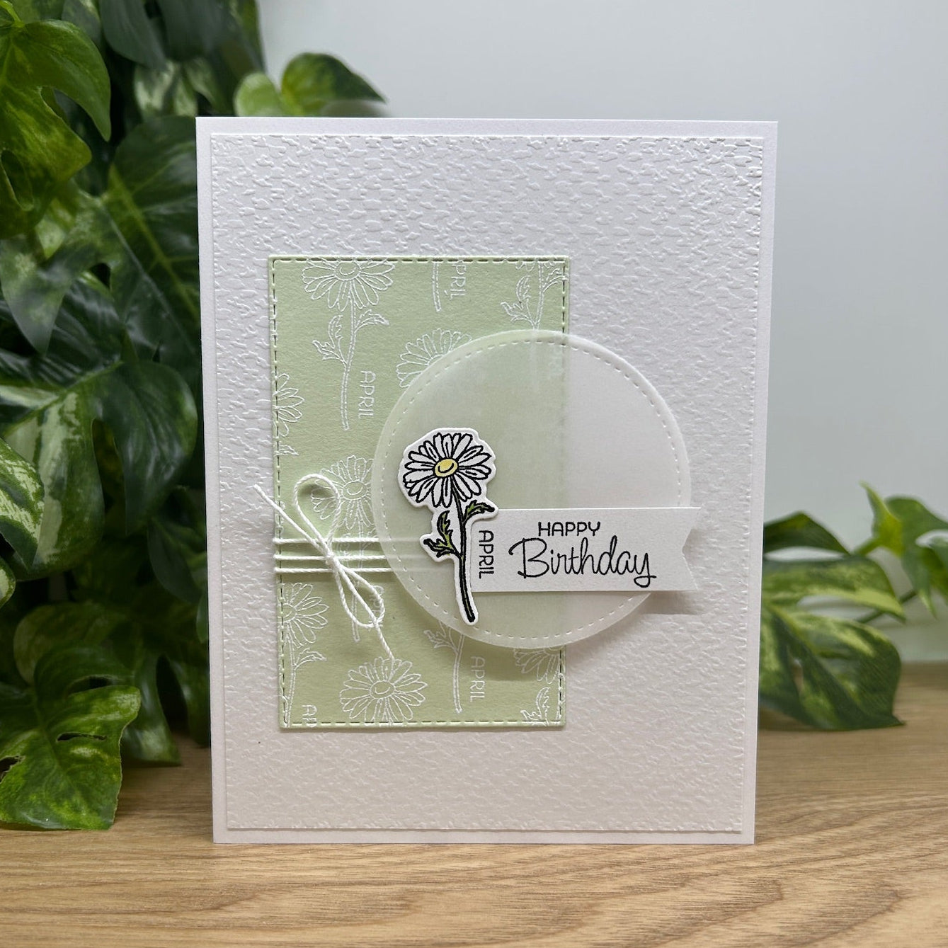 Handmade birthday card with floral design and 'Happy Birthday' text, placed on a wooden surface with green leaves in the background.