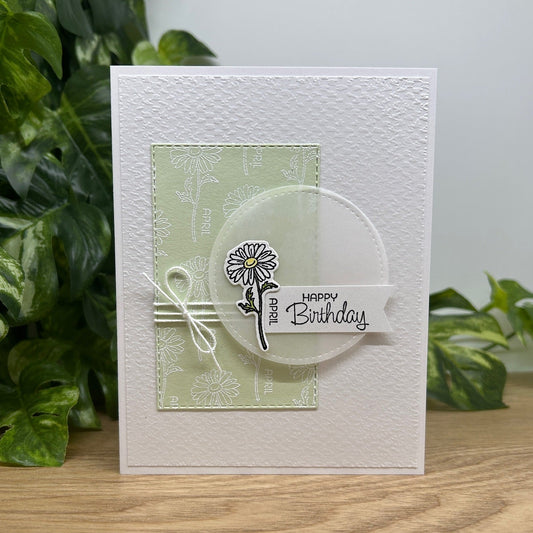 Handmade birthday card with floral design and 'Happy Birthday' text, placed on a wooden surface with green leaves in the background.