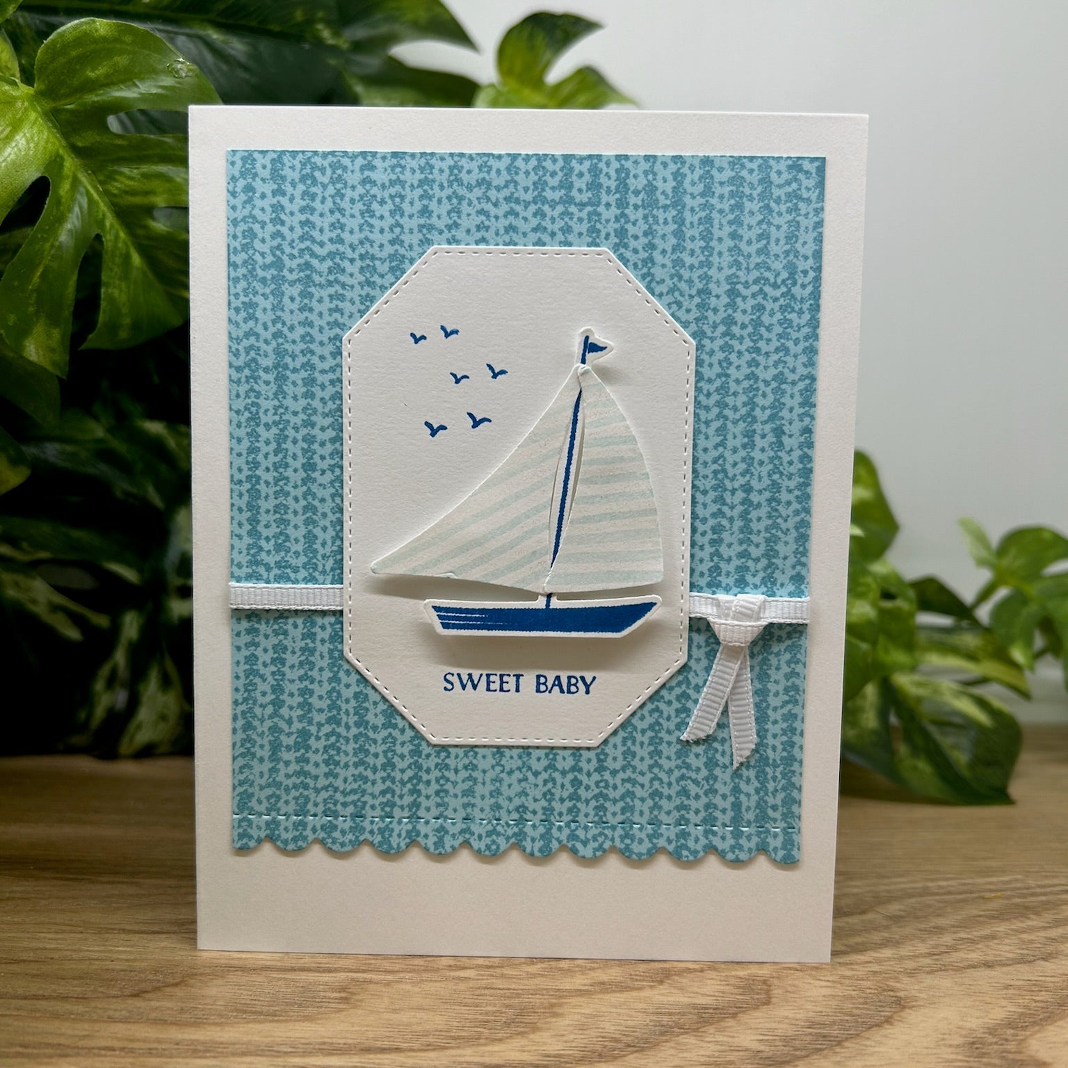 A handmade greeting card featuring a sailboat illustration with the text 'SWEET BABY' on it, presented on a wooden surface with a plant in the background.